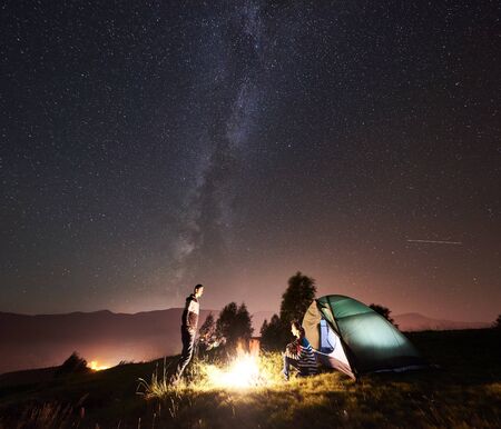 Happy romantic couple travelers resting at bonfire near glowing tourist tent under amazing night sky full of stars and Milky way. On the background starry sky, mountains and luminous cityの写真素材