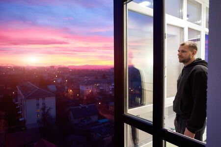 Side view of a man standing on a balcony with panoramic windows and looking into the distance while enjoying magical sunset. Urban landscape from height and a multi-colored fiery sky with setting sunの写真素材