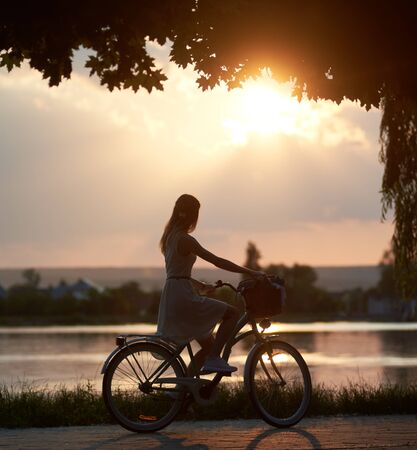 Tender girl rides a retro bike with a basket on the road near the river on a sunset. Girl enjoys the magical view of the suns rays of the falling sun at the end of the dayの写真素材
