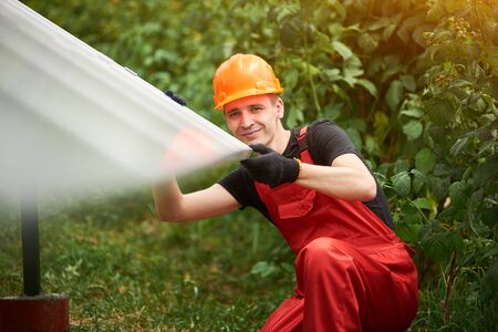 Happy man worker in orange uniform and safety helmet on blurry background of solar panels and greenery. Home construction of solar station. Alternative energy sustainable conceptの写真素材