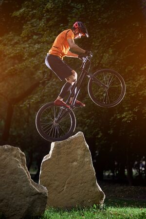 Extreme show of talented trail biker performing on big rocks standing on back wheel, in the evening in park, side view snapshot, concept of active lifestyleの写真素材