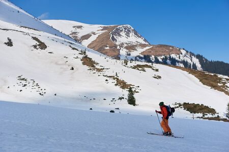 Skier tourist backpacker climbing on skis up steep hill on background of bright blue sky and...