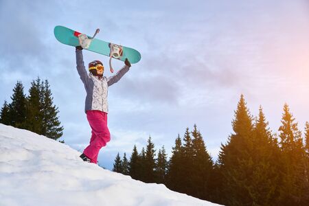 Sporty girl is standing on snowy hill holding her snowboard above her head, forest on background, copy space, concept of extreme winter sportの写真素材
