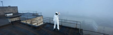 Panoramic snapshot of a scientist wearing white coverall and gas mask, standing on the roof of a high storey building with his hand up, gesturing stop sign in quarantineの写真素材