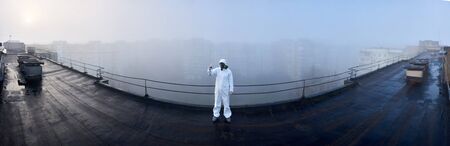 Panoramic snapshot of a man wearing white coverall and gas mask, standing on the roof of a high storey building, holding a glass flask with blue substance, making research on pollution studyの写真素材