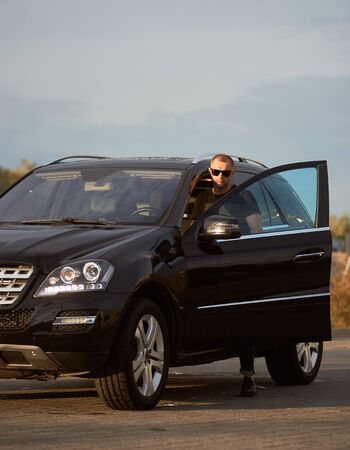 Portrait of handsome businessman wearing sunglasses is getting into his black car on the street under a blue skyの写真素材
