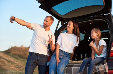 Father is taking selfie of his happy family sitting in the car trunk and having pizza and wine, family look white t-shirts and blue jeans. Man showing peace sign. Daughter showing thumbs upの写真素材