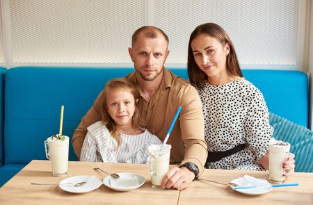 Family portrait of loving father is hugging his daughter while his wife is hugging him, smiling and looking in the camera, sitting in the cafe on blue sofa, milkshakes on the tableの写真素材