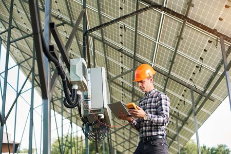 Serious man engineer checking the operation with laptop and sensor, on the background of the solar power station. Solar panels, sunny power electricity. Innovation future. Modern technology.の写真素材
