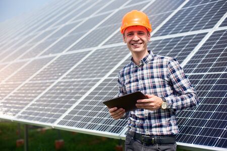 Portrait of smiling male engineer with tablet in his hands near the solar panels station, wearing helmet at sunny day. Green ecological power energy generation.の写真素材