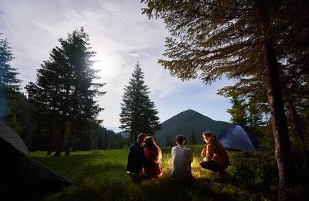 Loving couple and friends at night camping in the mountains. Tourist people sitting by campfire near tents under the spruce trees against the backdrop of the mountain valley.の写真素材