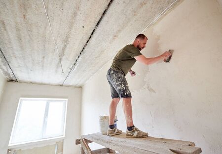 Apartment remodel. Worker is standing on wooden stand and puttying the walls indoors at small room. A guy with a beard in a t-shirt and jeans is smeared paintの写真素材