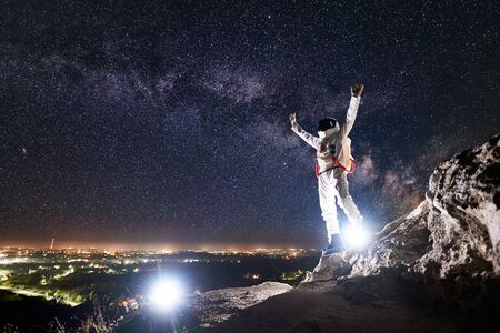 Spaceman raising arms while standing on rocky mountain with fantastic starry sky on background....