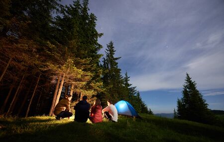 Night natural landscape and a group of tourists near a campfire and illuminated tourist tent. Mighty pine forest, evening sky with rare stars, valley of hills and mountains in the distanceの写真素材