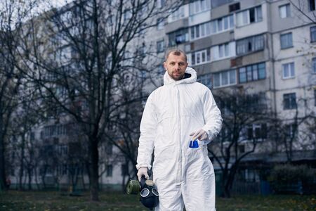 Portrait of a male scientist in white coverall, holding gas mask in one hand and glass flask in other, standing in front of a high residential building after examining the territoryの写真素材
