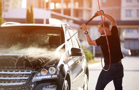 Side view of a man washing his car, holding a hose over his head, water jet under high pressure cleaning black car, surface of the vehicle is shining on the sunの写真素材