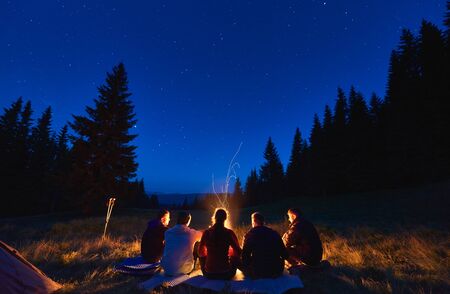 Summer camping under stars. Rear view of group of five hikers, men and woman sitting near bright bonfire, tourist tent under dark night sky with sparkling stars. Concept of tourism, night camping.の写真素材