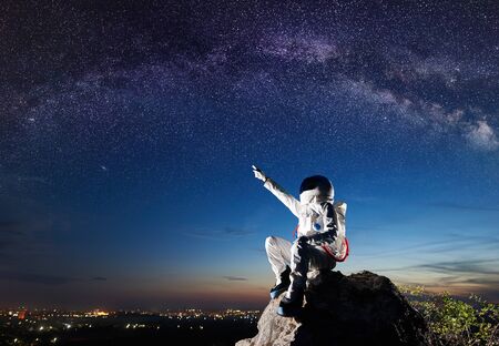Side view of spaceman pointing at beautiful sky with stars while sitting on top of mountain. Mission specialist astronaut wearing white space suit with helmet. Concept of space travel and night sky.の写真素材