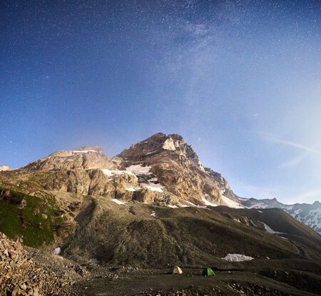 Amazing scenery in sunny summer day with blue sky and stars, beautiful mountains area with snow and green grass. Gorgeous mountain ridge with high rocky peaks, two colorful tents, wonderland.の写真素材