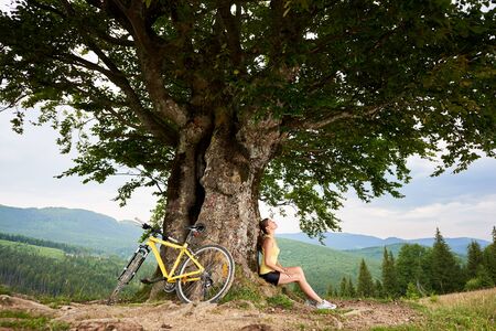 Attractive happy woman cyclist resting near yellow mountain bicycle under big tree, enjoying summer day in the mountains. Outdoor sport activity, lifestyle conceptの写真素材