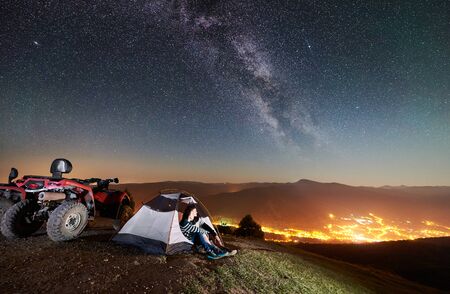 Young man and woman sitting in tourist tent on the top of mountain with atv quad motorbikes, romantic couple enjoying beautiful view of night sky full of stars, Milky way, luminous town on backgroundの写真素材