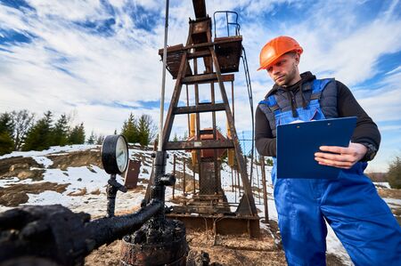 Serious oil man in work overalls and helmet holding clipboard and looking at oil pump rocker-machine. Male worker checking oil pumping unit, making notes. Concept of oil extraction, petroleum industryの写真素材