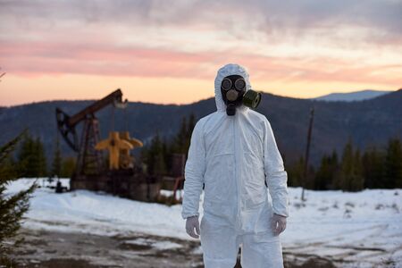 Scientist wearing white coverall, gloves and a gas mask working in oil field in mountainous region,...