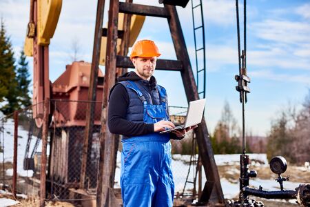 Portrait of petrolium engineer, wearing blue overalls and orange helmet, with a laptop, standing with his back to an oil rig, checking oil pumping unit, making notes in his computerの写真素材