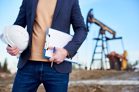 Cropped close-up of man engineer holding his note and pen in one hand and white helmet in the other, standing in oil field, oil pump jack is on background, concept of oil business, petroleum industryの写真素材