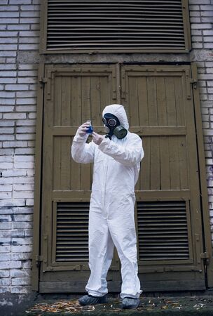 Full length snapshot of ecologist standing in front of old door and observing reaction in glass flask with blue liquid, laboratorial experiment. Scientist wearing white uniform, gloves and gas maskの写真素材