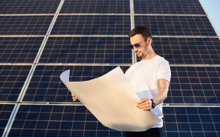 Businessman watching a plan on a background of solar panels. Young man with a clock in sunglasses, a white T-shirt and jeans familiarizes himself with an important documentの写真素材
