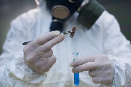 Cropped close-up snapshot of scientist's hands wearing gloves, holding a glass flask with blue liquid and a tweezers with a cigarette butt as a sample.の写真素材