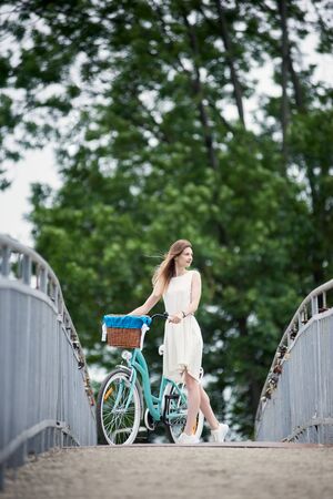 Cheerful maiden in a light dress with long hair standing with a vintage blue bike and enjoing nature on the bridge that leads to the park with green trees. Blurred backgroundの写真素材