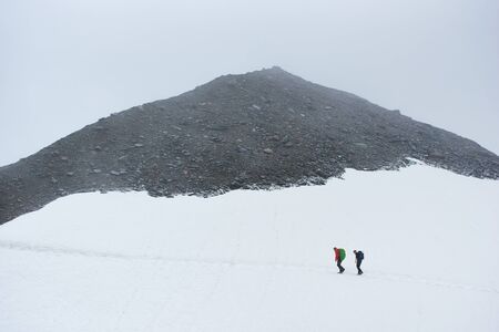 Beautiful view of winter rocky mountains with two male travelers. Mountaineers with backpacks walking through the snow with rocky hill on background. Concept of travelling, mountaineering and hiking.の写真素材