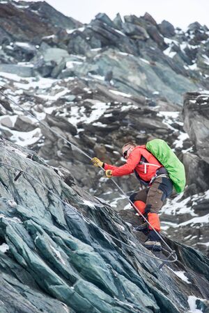 Brave mountaineer in sunglasses holding rope while climbing alpine ridge. Male alpinist with backpack ascending mountain and trying to reach mountaintop. Concept of alpinism and alpine climbing.の写真素材