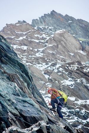 Mountaineer with backpack using fixed rope to climb high rocky mountain. Man climber ascending alpine ridge and trying to reach mountaintop. Concept of mountaineering and alpine rock climbing.の写真素材