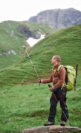 Full length of traveler backpacker pointing with trekking poles. Young man standing on rock with grassy hills while traveling alone in mountains. Concept of hiking and active lifestyle.の写真素材