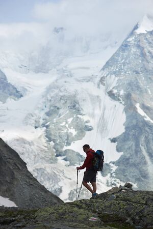 Tourist with backpack walking using trekking sticks, beautiful mountains with snow. Mountain hiking, man reaching peak. Wild nature with amazing views. Sport tourism in Alps.の写真素材