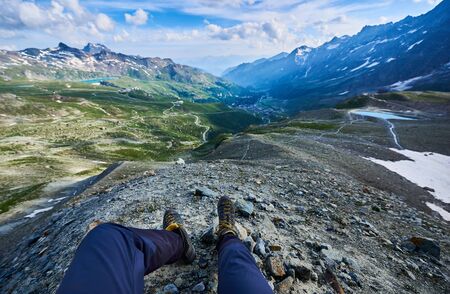 Male legs in hiking boots resting on hillside in mountain valley. Beautiful view of mountains and hills under cloudy sky with man hiker feet on foreground. Concept of travelling, hiking and alpinism.の写真素材