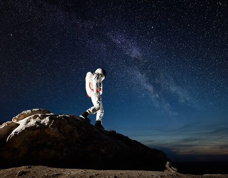 Astronaut walking down the rocky hill under fantastic night sky with stars. Cosmonaut in space suit...