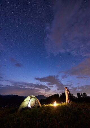 Male tourist spending time in the mountains in the evening with illuminated tent and a campfire under beautiful sky full of stars. Night camping in the mountains at summerの写真素材