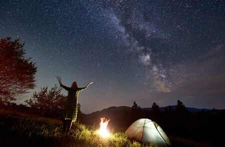 Amazing starry night in the mountains, girl is standing with her hands up and looking at Milky Way constellation, near camping place with a tent and a fireの写真素材
