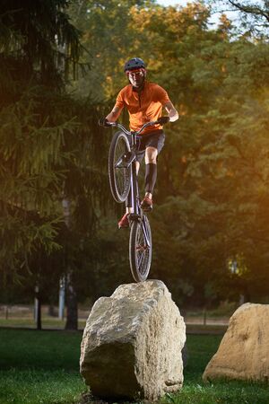 Portrait of young cyclist standing on back wheel of bmx bike on a rock looking at camera in park, green trees on backgroundの写真素材