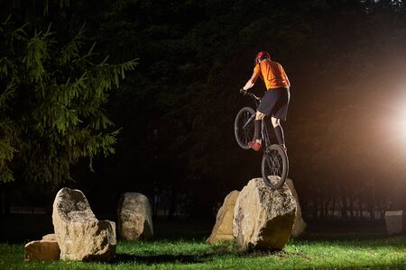 Extreme cyclist with his back to camera on bmx bike in park performing stunt keeping balance on back wheel on rock, in the evening, copy spaceの写真素材