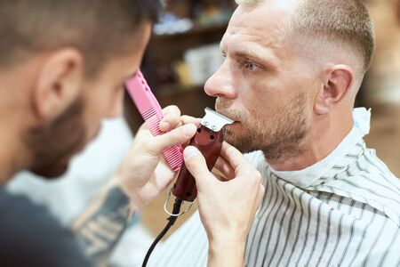 Close-up side view of a cool man at the barber shop having his mustache cut with a red trimmer, blurred side view of a bearded hairdresser holding toolsの写真素材