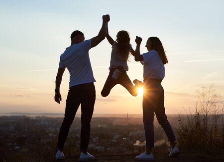 Silhouette of father mother and their daughter spending fun day outside the city on the hill on the sunset with a beautiful city view, dad and mom holding girl by hands and she is hanging, back view