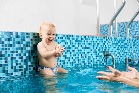Cute playful baby boy sitting on a silver pipe in the swimming pool preparing to jump in water to his moms hands. Concept of healthy chindhoodの写真素材