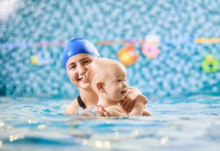 Swimming class for infants. Young smiling mother supporting her baby in swimming pool. Happy little boy enjoying interaction with his mom. Concept of happy childhoodの写真素材
