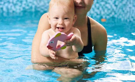 Cropped close-up snapshot of a cheerful playful little boy in his mothers embrace in turquoise water in swimming pool. Baby is showing his toy fish to the camera. Happy family conceptの写真素材