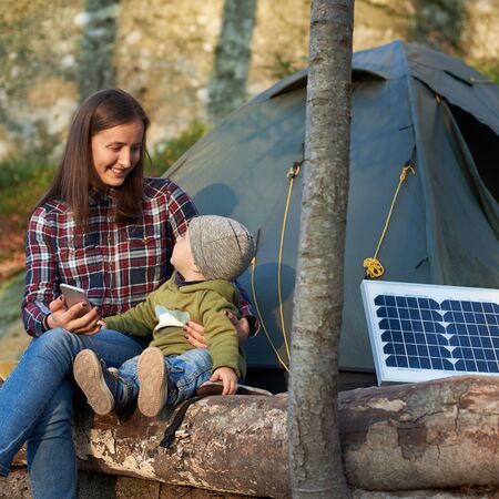 Woman looking at a child, smiling, sitting on a log in the forest near small solar panel and a tent camp. Girl is holding a telephone. Family weekend with the development of new technologiesの写真素材